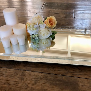 May include: A mirrored tray with a decorative etched border, featuring a bouquet of white and yellow flowers and four white candles.