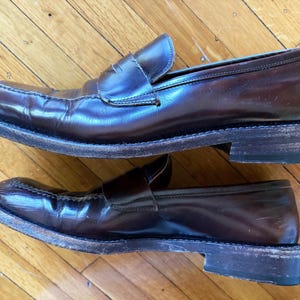 May include: A pair of dark brown leather penny loafers featuring a classic slip-on design with a strap across the vamp, a rounded toe, and a stacked heel. The shoes have visible signs of wear on the leather uppers and the edges of the soles.