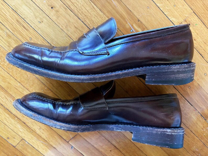 May include: A pair of dark brown leather penny loafers featuring a classic slip-on design with a strap across the vamp, a rounded toe, and a stacked heel. The shoes have visible signs of wear on the leather uppers and the edges of the soles.