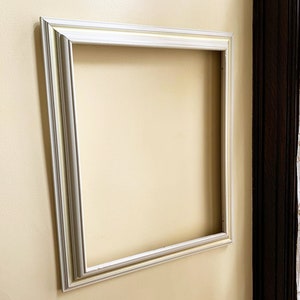 May include: A white picture frame with a simple, ornate design. The frame is mounted on a light yellow wall.
