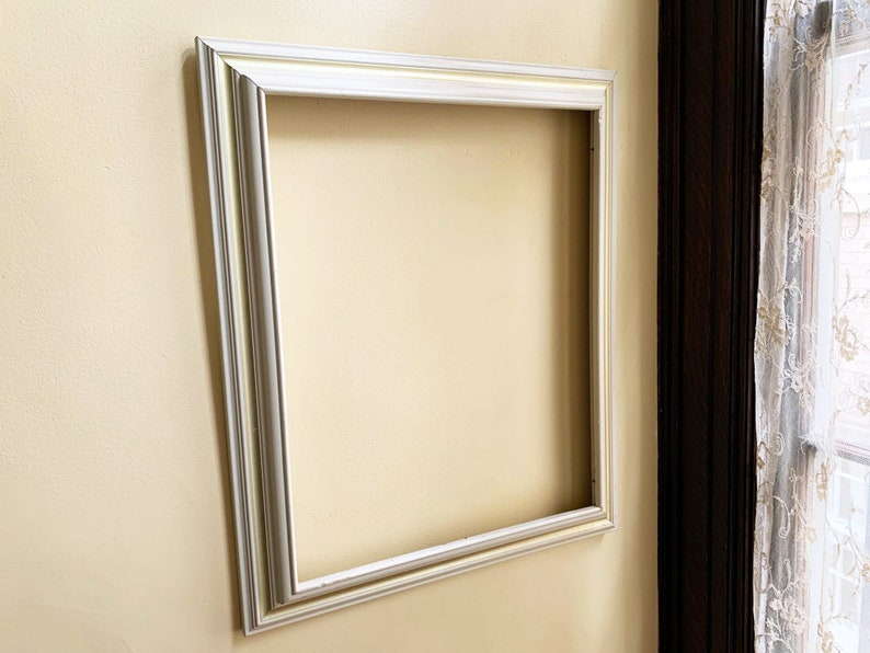 May include: A white picture frame with a simple, ornate design. The frame is mounted on a light yellow wall.