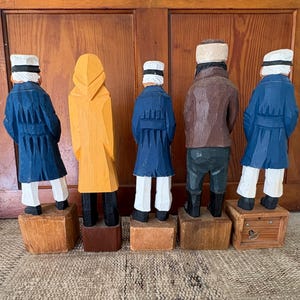 May include: Five carved wooden figures of sailors, viewed from the back. The figures are painted in various colors, including blue, yellow, and brown. Each figure stands on a wooden block. The figures are wearing hats and coats.