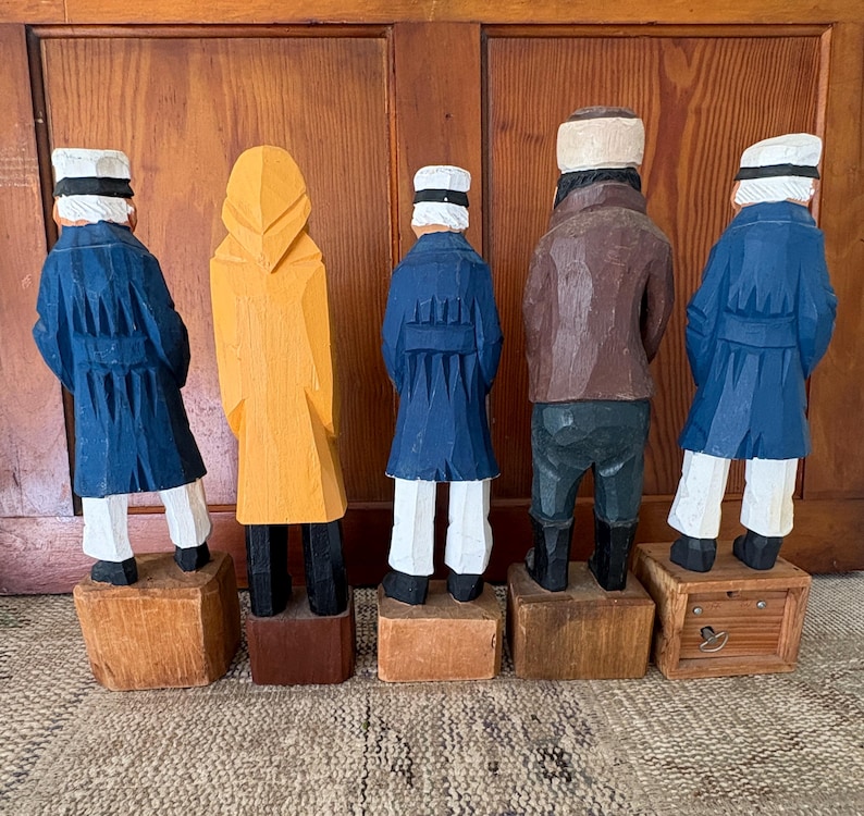 May include: Five carved wooden figures of sailors, viewed from the back. The figures are painted in various colors, including blue, yellow, and brown. Each figure stands on a wooden block. The figures are wearing hats and coats.