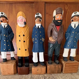 May include: Five hand-carved wooden sailor figurines. The figures are dressed in blue, yellow, and brown coats, white pants, and black boots. Each figure stands on a small wooden block. The sailor figurines have painted faces and hats.