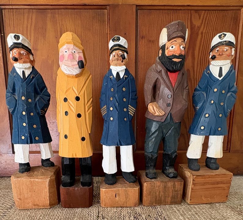 May include: Five hand-carved wooden sailor figurines. The figures are dressed in blue, yellow, and brown coats, white pants, and black boots. Each figure stands on a small wooden block. The sailor figurines have painted faces and hats.