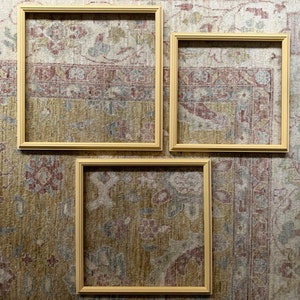 May include: Three empty picture frames with a light wood finish, arranged in a row on a patterned rug. The frames are all the same size and shape, and they are all oriented horizontally.