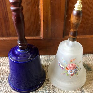 May include: Two decorative bells with wooden handles. One bell is blue with a gold trim and the other is white with a floral design.