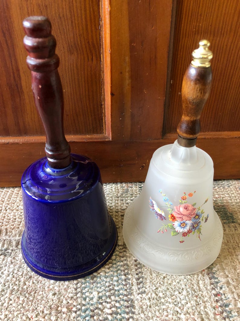 May include: Two decorative bells with wooden handles. One bell is blue with a gold trim and the other is white with a floral design.