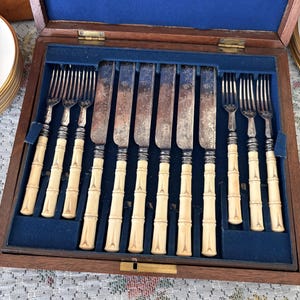 Pode incluir: Um conjunto de talheres antigos, incluindo garfos e facas, exibido numa caixa de madeira com forro de veludo azul. As pegas t&ecirc;m um design de bambu e as l&acirc;minas apresentam gravuras decorativas.