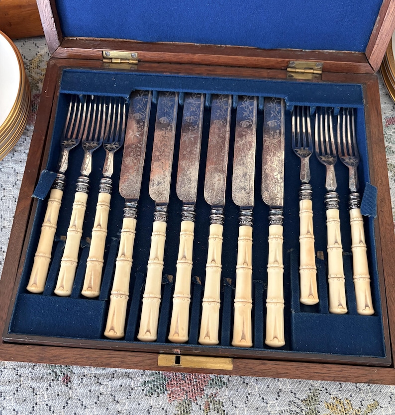 Pode incluir: Um conjunto de talheres antigos, incluindo garfos e facas, exibido numa caixa de madeira com forro de veludo azul. As pegas t&ecirc;m um design de bambu e as l&acirc;minas apresentam gravuras decorativas.