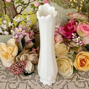 Peut inclure: Un vase blanc de style vintage avec un bord festonn&eacute; et un corps facett&eacute;. Le vase est entour&eacute; de fleurs color&eacute;es, dont des roses roses, jaunes et p&ecirc;che, sur fond d'&oelig;uvre florale brod&eacute;e.