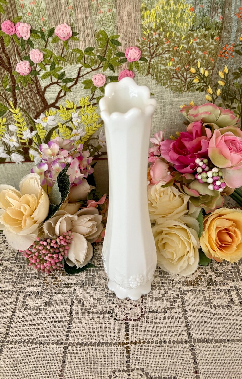 Peut inclure: Un vase blanc de style vintage avec un bord festonn&eacute; et un corps facett&eacute;. Le vase est entour&eacute; de fleurs color&eacute;es, dont des roses roses, jaunes et p&ecirc;che, sur fond d'&oelig;uvre florale brod&eacute;e.