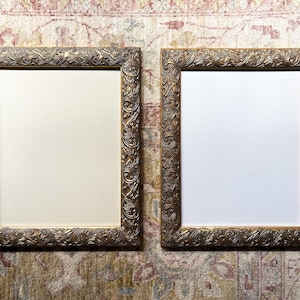 May include: Two ornate, rectangular picture frames with detailed gold-colored scrollwork. One frame has a cream-colored interior, the other has a white interior. The frames are displayed on a patterned rug.
