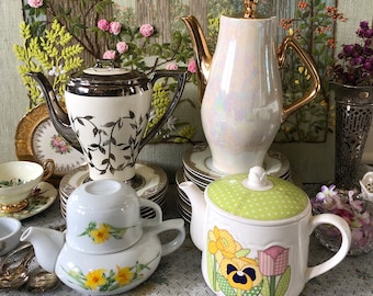 May include: A collection of four teapots in various styles and colors. The teapots are arranged on a white tablecloth with a floral pattern. The teapots are made of ceramic and have different designs, including floral patterns, polka dots, and a silver-leaf design.