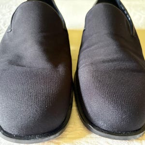 Puede incluir: Un par de zapatos negros sin cordones. Los zapatos tienen una parte superior de tela texturizada y una suela negra. El dise&ntilde;o es simple y cl&aacute;sico, adecuado para diversas ocasiones. Los zapatos se muestran sobre una superficie de color claro.
