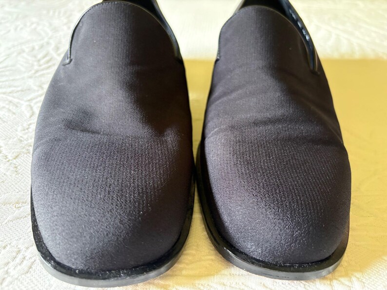 Puede incluir: Un par de zapatos negros sin cordones. Los zapatos tienen una parte superior de tela texturizada y una suela negra. El dise&ntilde;o es simple y cl&aacute;sico, adecuado para diversas ocasiones. Los zapatos se muestran sobre una superficie de color claro.
