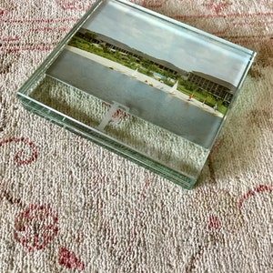 May include: A glass picture frame with a photo of a beach scene with sailboats. The frame is sitting on a white and pink textured surface.