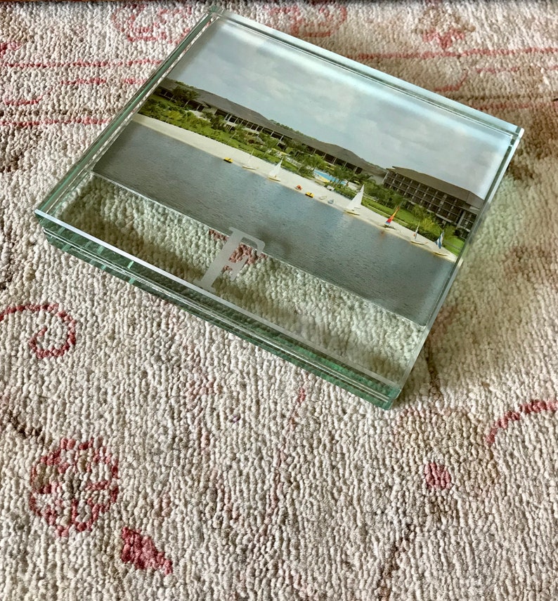 May include: A glass picture frame with a photo of a beach scene with sailboats. The frame is sitting on a white and pink textured surface.