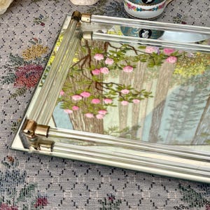 May include: A rectangular mirrored tray with a forest scene featuring pink flowers and trees. The tray has a clear glass handle with gold-colored metal accents. The tray is on a floral patterned surface.