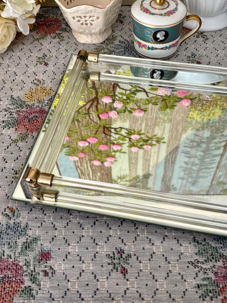 May include: A rectangular mirrored tray with a forest scene featuring pink flowers and trees. The tray has a clear glass handle with gold-colored metal accents. The tray is on a floral patterned surface.