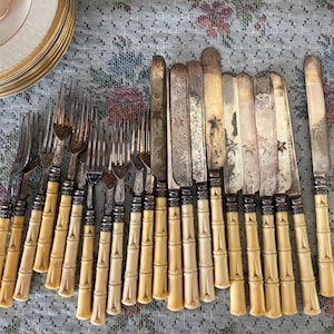 Pode incluir: Uma cole&ccedil;&atilde;o de talheres antigos, incluindo garfos e facas, com cabos em estilo bambu. Os talheres est&atilde;o dispostos sobre uma toalha de mesa com padr&atilde;o floral, com uma pilha de pratos com borda dourada ao fundo.