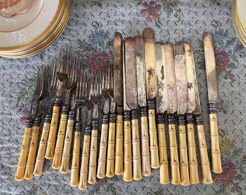 Pode incluir: Uma cole&ccedil;&atilde;o de talheres antigos, incluindo garfos e facas, com cabos em estilo bambu. Os talheres est&atilde;o dispostos sobre uma toalha de mesa com padr&atilde;o floral, com uma pilha de pratos com borda dourada ao fundo.