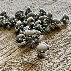 May include: A group of 17 silver-toned round cabinet knobs with a smooth, shiny finish. The knobs are arranged in a pile on a beige textured surface.