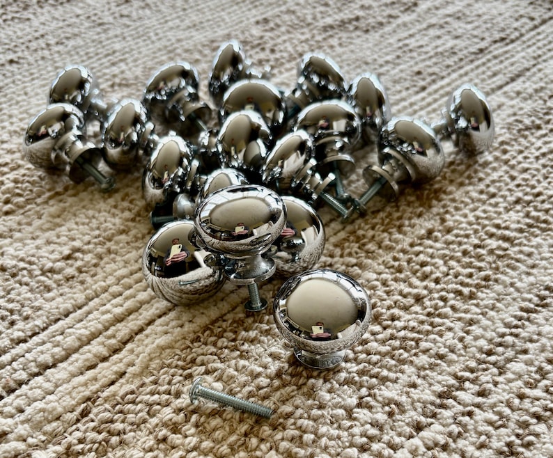 May include: A group of 17 silver-toned round cabinet knobs with a smooth, shiny finish. The knobs are arranged in a pile on a beige textured surface.