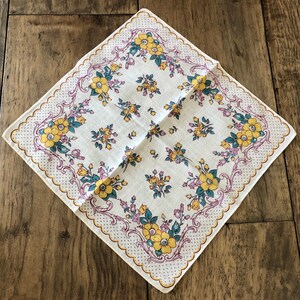 K&ouml;nnte beinhalten: Quadratisches wei&szlig;es Taschentuch mit floralem Muster. Das Taschentuch hat gelbe und t&uuml;rkisfarbene Blumen mit lila Akzenten. Der Rand hat einen gewellten Rand mit einem gepunkteten Design. Das Taschentuch liegt auf einer Holzoberfl&auml;che.