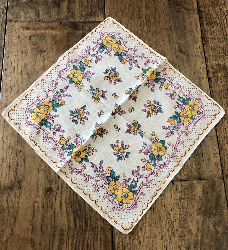 K&ouml;nnte beinhalten: Quadratisches wei&szlig;es Taschentuch mit floralem Muster. Das Taschentuch hat gelbe und t&uuml;rkisfarbene Blumen mit lila Akzenten. Der Rand hat einen gewellten Rand mit einem gepunkteten Design. Das Taschentuch liegt auf einer Holzoberfl&auml;che.