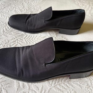 Puede incluir: Un par de zapatos negros sin cordones. Los zapatos tienen una punta redondeada y un tac&oacute;n bajo. La parte superior est&aacute; hecha de una tela lisa y oscura. Los zapatos se exhiben sobre una superficie blanca y texturizada.