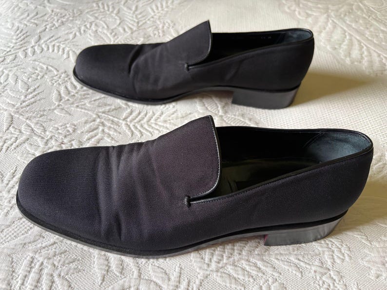 Puede incluir: Un par de zapatos negros sin cordones. Los zapatos tienen una punta redondeada y un tac&oacute;n bajo. La parte superior est&aacute; hecha de una tela lisa y oscura. Los zapatos se exhiben sobre una superficie blanca y texturizada.