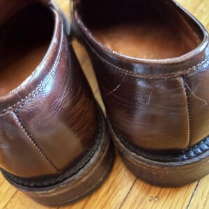 May include: A pair of dark brown leather loafers featuring a polished finish, visible stitching along the heel counter, and a stacked heel with a textured welt. The interior lining is a lighter tan leather, and the exterior shows natural creasing.