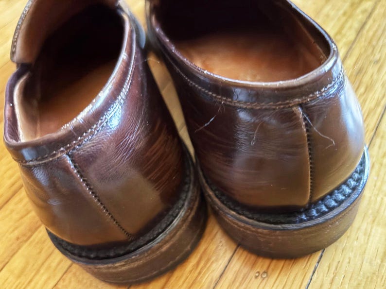 May include: A pair of dark brown leather loafers featuring a polished finish, visible stitching along the heel counter, and a stacked heel with a textured welt. The interior lining is a lighter tan leather, and the exterior shows natural creasing.