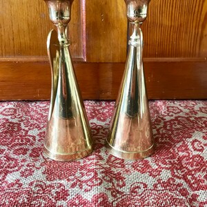 May include: A pair of gold metal candle holders with a conical shape and a decorative detail on the side. The candle holders are on a red and white patterned surface.