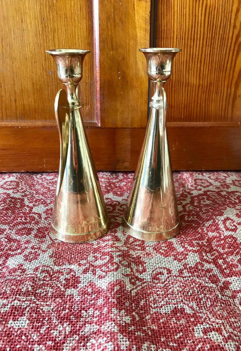 May include: A pair of gold metal candle holders with a conical shape and a decorative detail on the side. The candle holders are on a red and white patterned surface.
