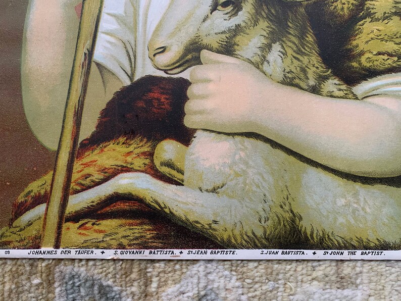 May include: A vintage religious image depicting a young boy holding a lamb in his arms. The boy is wearing a white tunic and has a halo above his head. The image is titled "St. John the Baptist" in English, German, Italian, Spanish, and Portuguese.