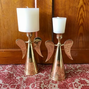 May include: Two gold angel candle holders with white candles. The candle holders are made of metal and have a cone-shaped base with wings.