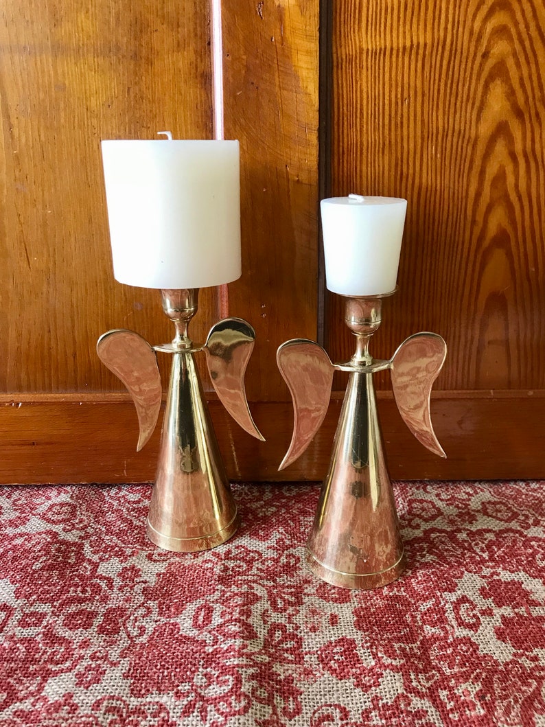 May include: Two gold angel candle holders with white candles. The candle holders are made of metal and have a cone-shaped base with wings.