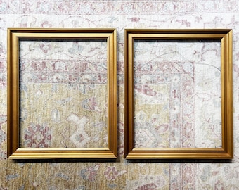 May include: Two rectangular, empty gold picture frames. The frames are made of wood and have a simple, classic design. The frames are displayed on a patterned rug with a mix of colors.