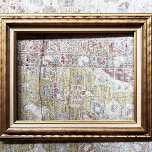 May include: A rectangular picture frame with a metallic gold finish and a stepped profile. The frame features a light beige inner border and a thin, dark vertical wire hanging down from the top inner edge, centred within the open space.