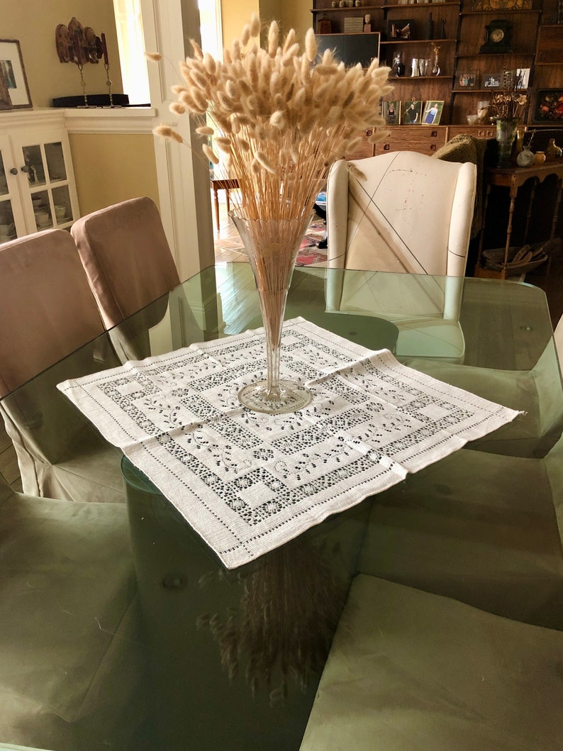 Peut inclure: Une table en verre avec une nappe blanche et un vase de pampas s&eacute;ch&eacute;es au centre. La table est entour&eacute;e de chaises avec des coussins beiges.