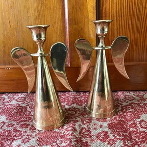 May include: Two gold angel candle holders with wings and a cone-shaped base. The candle holders are made of metal and have a polished finish.