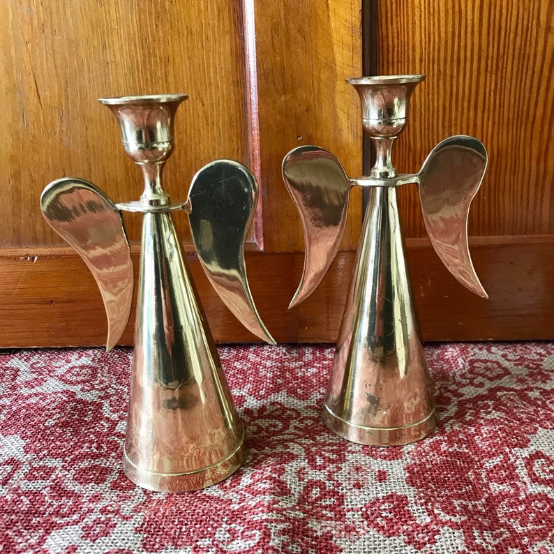 May include: Two gold angel candle holders with wings and a cone-shaped base. The candle holders are made of metal and have a polished finish.