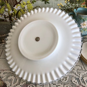 Puede incluir: Un soporte para tartas de vidrio blanco lechoso con un borde festoneado. El soporte tiene un pedestal central y una superficie lisa y blanca.
