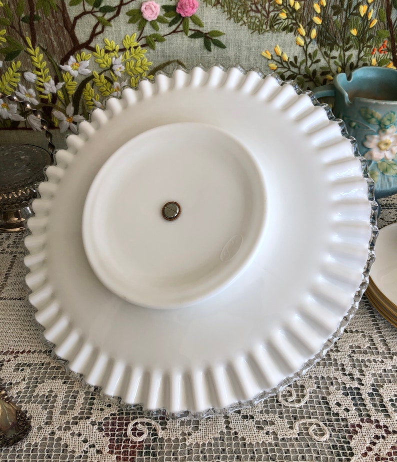 Puede incluir: Un soporte para tartas de vidrio blanco lechoso con un borde festoneado. El soporte tiene un pedestal central y una superficie lisa y blanca.