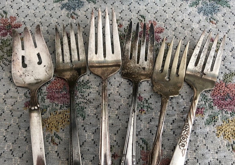 K&ouml;nnte beinhalten: Sieben silberne Gabeln mit unterschiedlichen Designs und Zinken. Die Gabeln sind in einer Reihe auf einer gemusterten Tischdecke angeordnet.