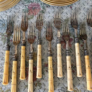 Pode incluir: Um conjunto de garfos antigos com cabos em estilo bambu e dentes prateados. Os cabos s&atilde;o de cor creme claro com detalhes prateados. Os garfos est&atilde;o dispostos sobre uma toalha de mesa com padr&atilde;o floral, com pratos dourados ao fundo.