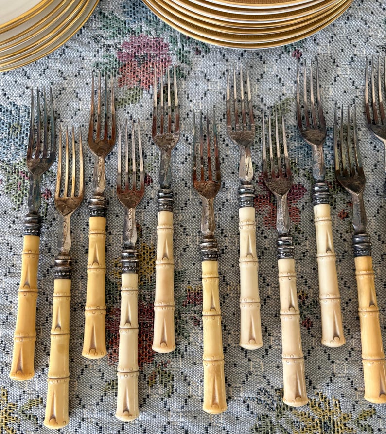 Pode incluir: Um conjunto de garfos antigos com cabos em estilo bambu e dentes prateados. Os cabos s&atilde;o de cor creme claro com detalhes prateados. Os garfos est&atilde;o dispostos sobre uma toalha de mesa com padr&atilde;o floral, com pratos dourados ao fundo.