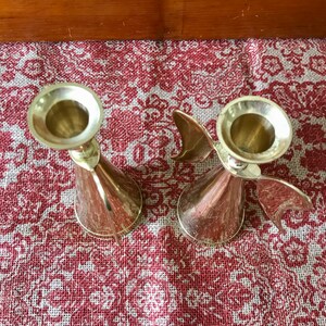 May include: Two gold metal candle holders with a wing design. The candle holders are on a red and white patterned surface.